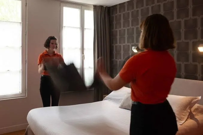 Mitarbeiterin des Hotels Victoria in orangefarbenem Poloshirt beim Bettenmachen in einem in Grautönen gehaltenen Zimmer