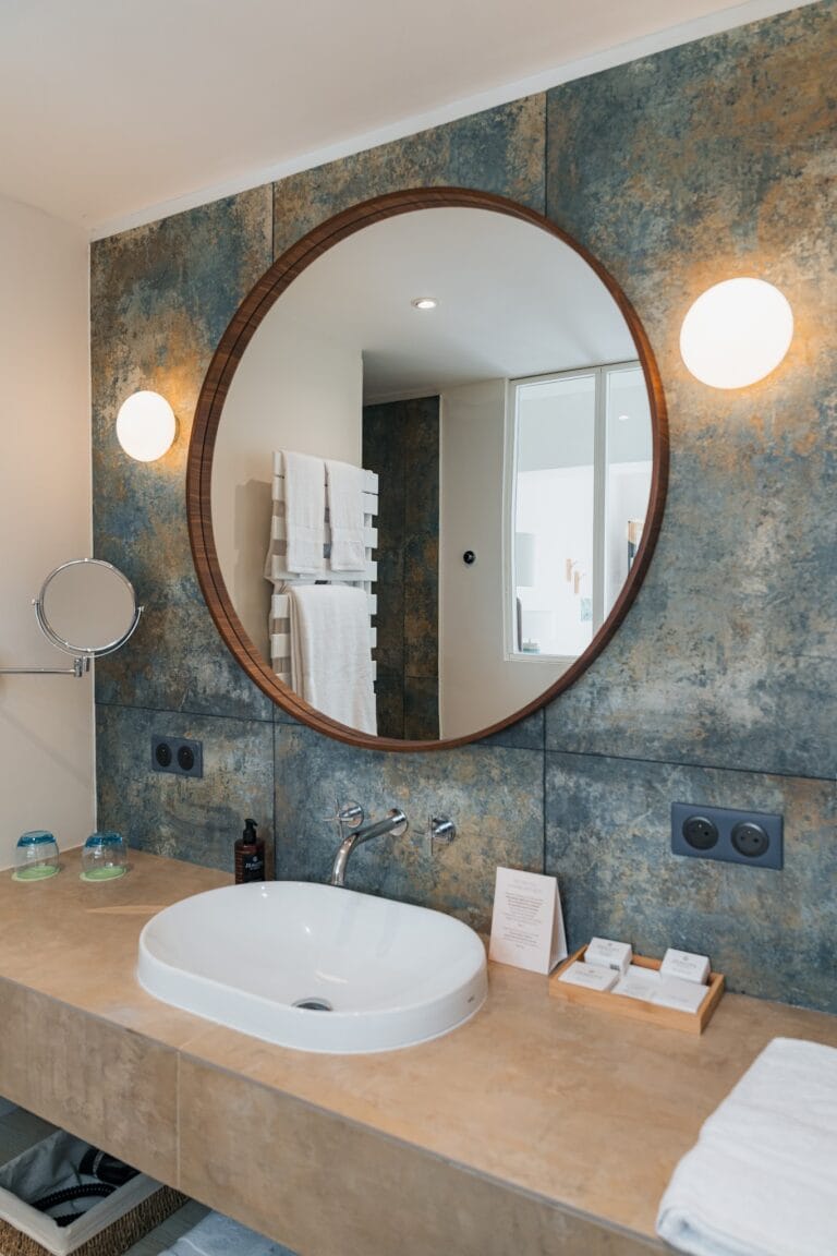 Bathroom at l'Esquisse Hôtel Culturel, oval white washbasin and large round mirror on blue concrete-effect tiles