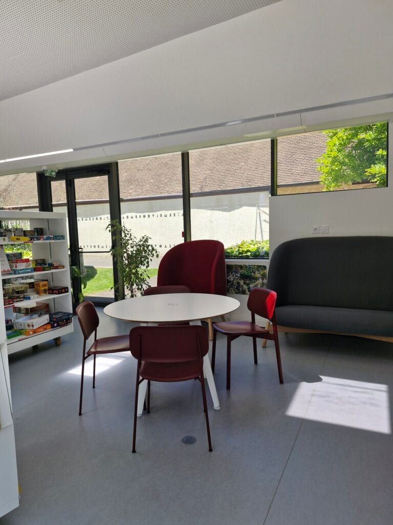 A convivial reading area at the Les Sources Vives multimedia library in Avon, with burgundy armchairs and a round table with a garden view.